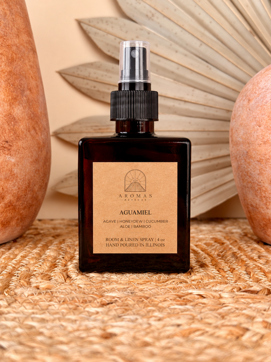 Aromas Aztecas Aguamiel room and linen spray in an amber glass bottle on a woven textured surface with terracotta pottery and dried palm leaf décor.