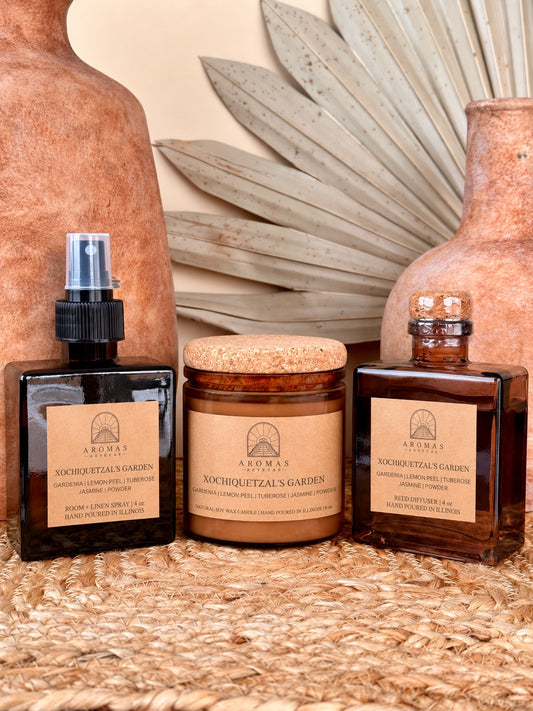 Aromas Aztecas Xochiquetzal's Garden room and linen spray, soy candle, and reed diffuser in amber glass vessels on a woven textured surface with terracotta pottery and dried palm leaf décor.