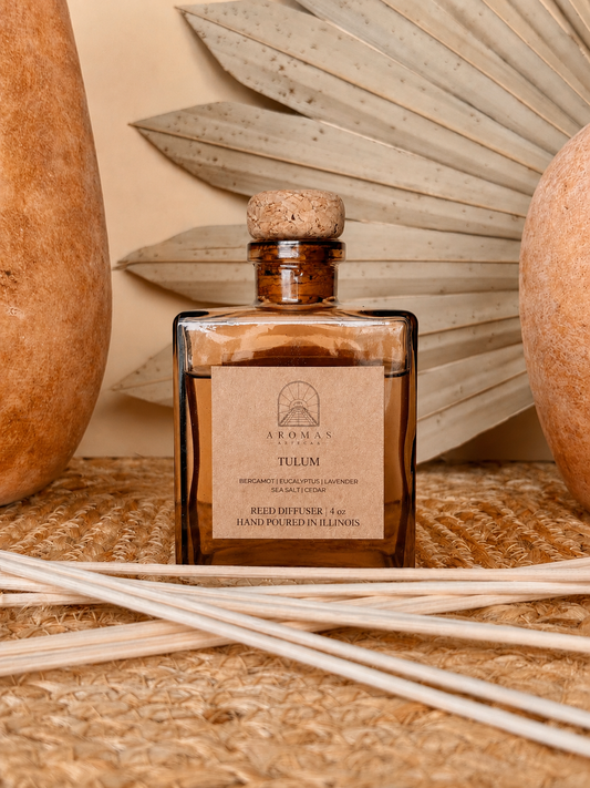 Aromas Aztecas Tulum reed diffuser in an amber glass bottle with natural reeds on a woven textured surface with terracotta pottery and dried palm leaf décor.