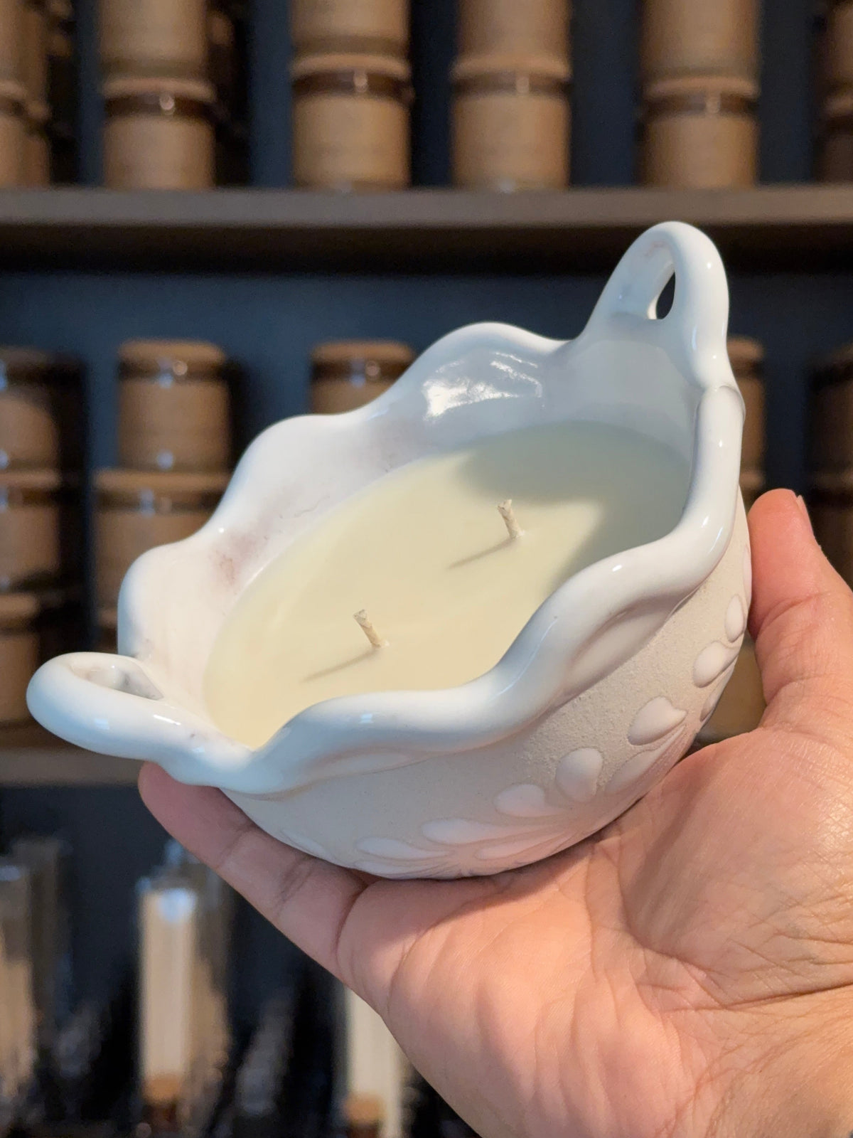 Handcrafted white talavera cazuelita candle with two wicks, held in a hand against a softly blurred shelf of candle vessels.