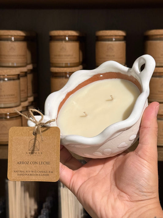 Arroz con Leche cazuelita candle with two wicks in a white artisan talavera vessel, held in a hand with a branded product tag, photographed against shelves of candle jars.