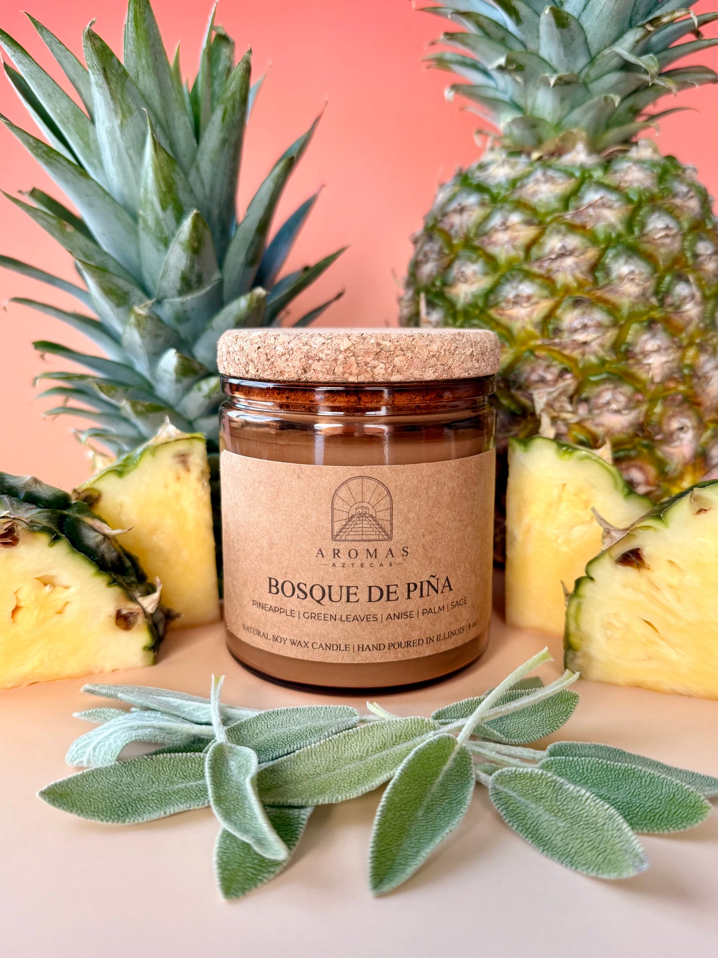Aromas Aztecas Bosque de Piña soy candle in an amber glass vessel with a cork lid on a sunset pink background surrounded by pineapple and sage.