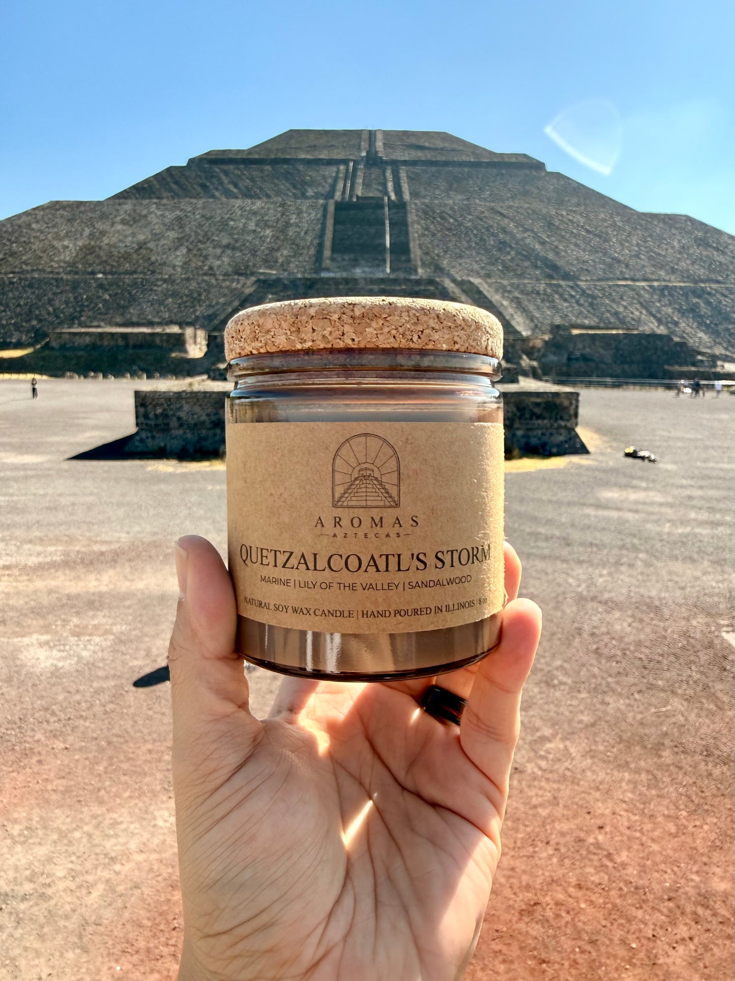 Aromas Aztecas Quetzalcoatl's Storm soy candle in an amber glass vessel with a cork lid held in front of the Pyramid of the Sun in Teotihuacán, México.