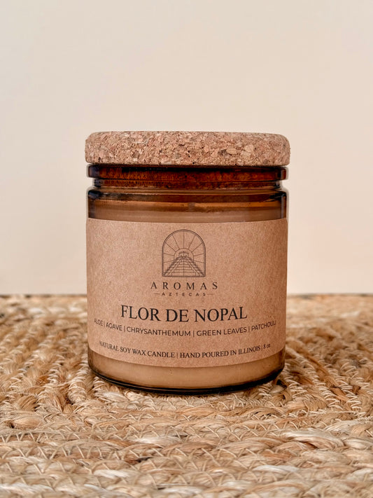 Aromas Aztecas Flor de Nopal soy candle in an amber glass vessel with a cork lid on a woven textured surface.