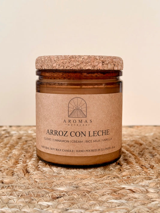 Aromas Aztecas Arroz con Leche soy candle in an amber glass vessel with a cork lid on a woven textured surface.