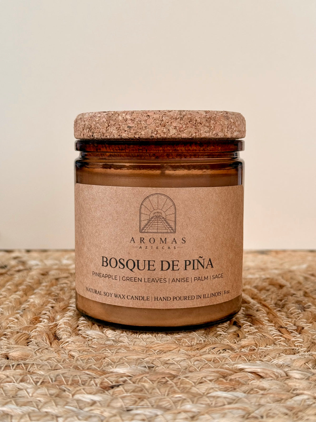 Aromas Aztecas Bosque de Piña soy candle in an amber glass vessel with a cork lid on a woven textured surface.