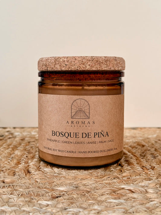 Aromas Aztecas Bosque de Piña soy candle in an amber glass vessel with a cork lid on a woven textured surface.