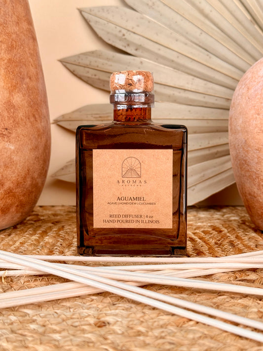 Aromas Aztecas Aguamiel reed diffuser in an amber glass bottle with natural reeds on a woven textured surface with terracotta pottery and dried palm leaf décor.