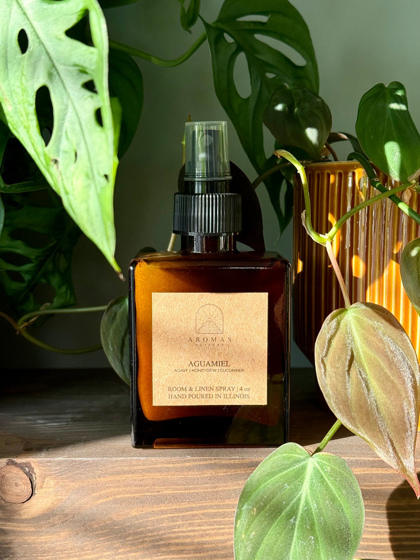Aromas Aztecas Aguamiel room and linen spray in an amber glass bottle on a wooden textured surface next to an deep orange plant pot and surrounded by leaves.
