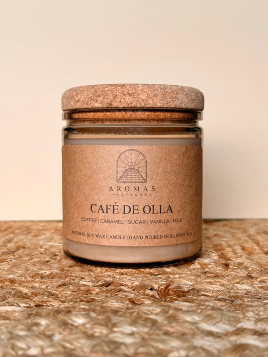 Aromas Aztecas Café de Olla soy candle in an amber glass vessel with a cork lid on a woven textured surface.