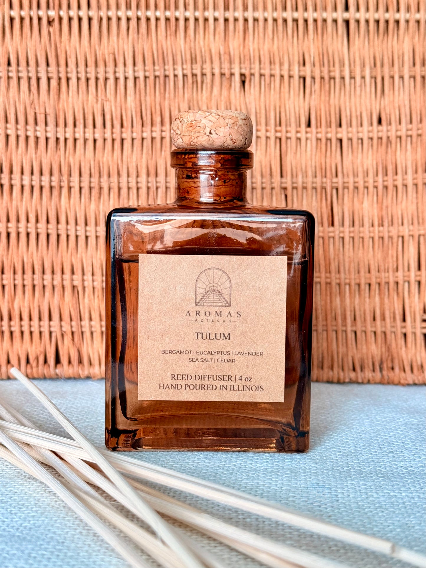Aromas Aztecas Tulum reed diffuser in an amber glass bottle with natural reeds against a woven textured backdrop.