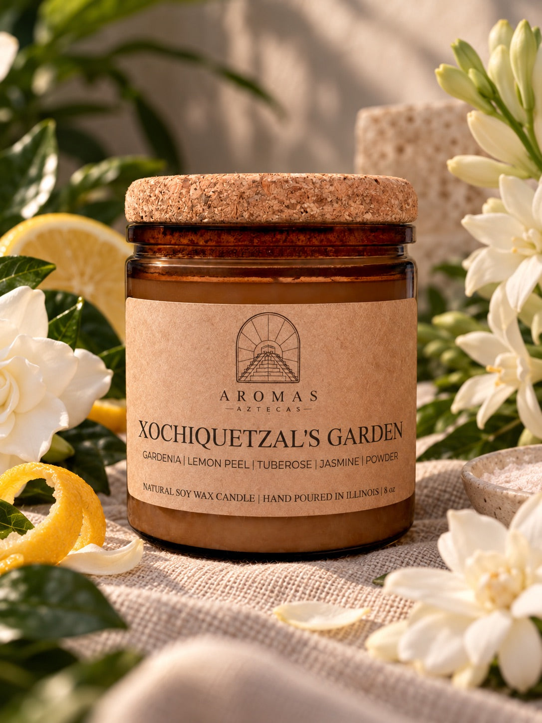 Aromas Aztecas Xochiquetzal’s Garden candle in an amber glass jar with a cork lid, displayed on neutral fabric with white gardenia and tuberose flowers, green leaves, and fresh lemon peel. 