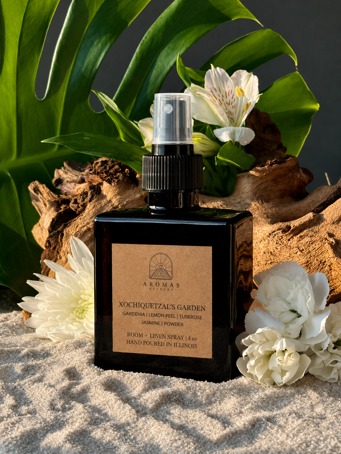 Aromas Aztecas Xochiquetzal's Garden room and linen spray in an amber glass bottle on a sandy surface with wood, flowers and leaves as a backdrop.