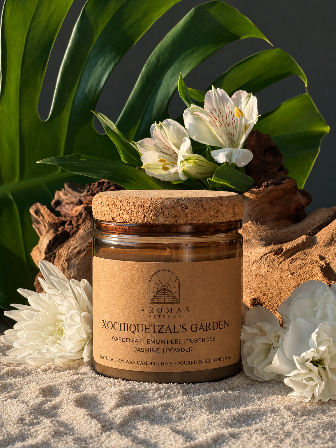 Aromas Aztecas Xochiquetzal's Garden soy candle in an amber glass vessel with a cork lid on a sandy surface and surrounded by wood, leaves, and flowers.