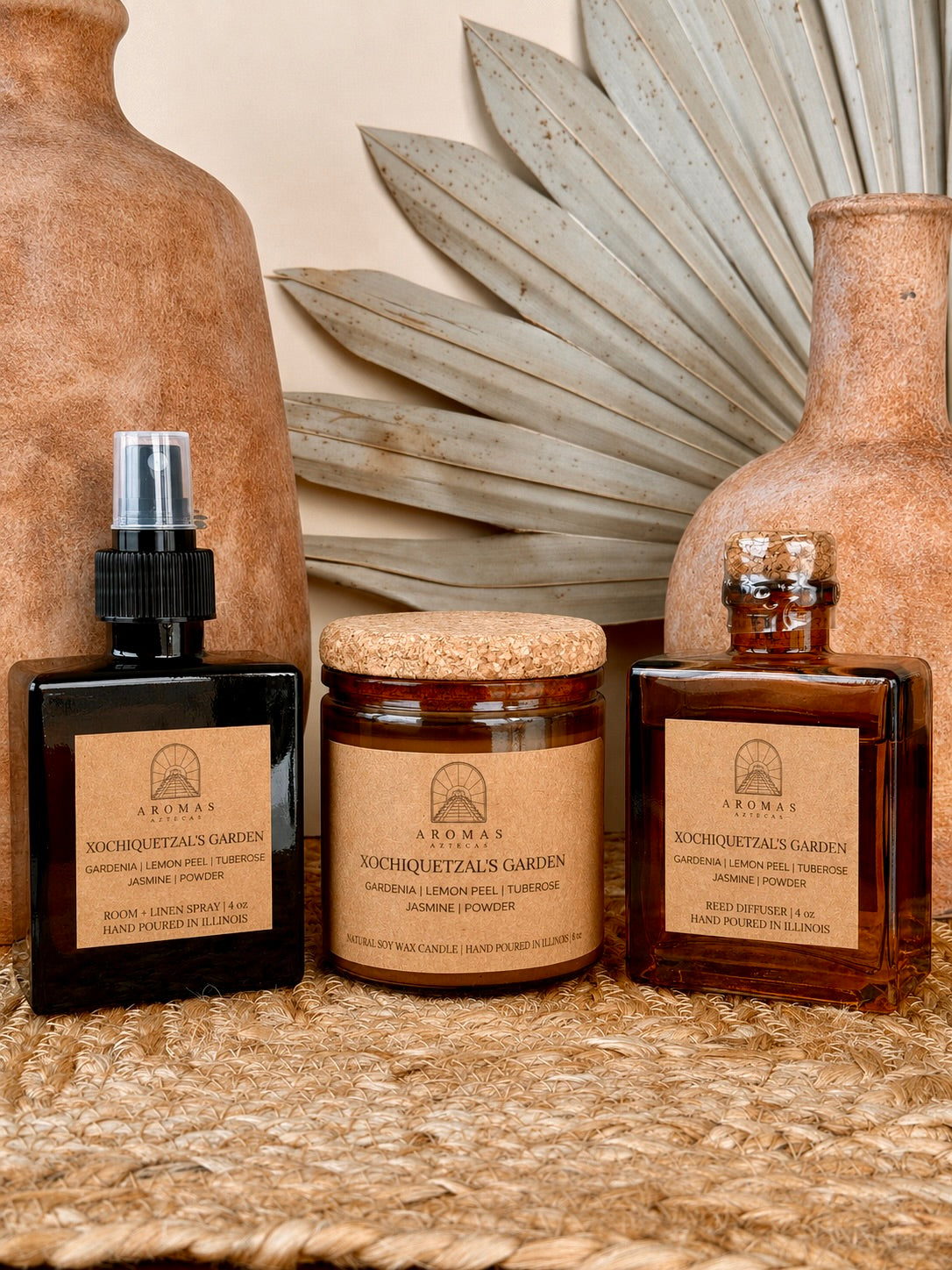 Aromas Aztecas Xochiquetzal's Garden room and linen spray, soy candle, and reed diffuser in amber glass vessels on a woven textured surface with terracotta pottery and dried palm leaf décor.