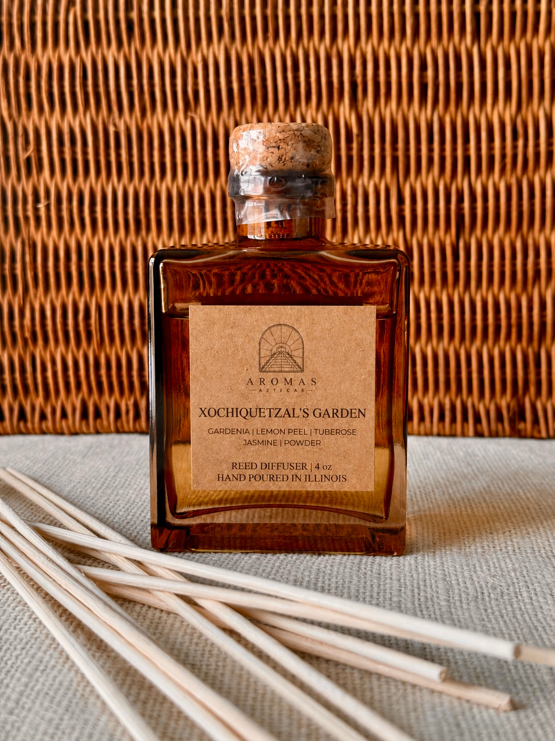 Aromas Aztecas Xochiquetzal's Garden reed diffuser in an amber glass bottle with natural reeds against a woven textured backdrop.