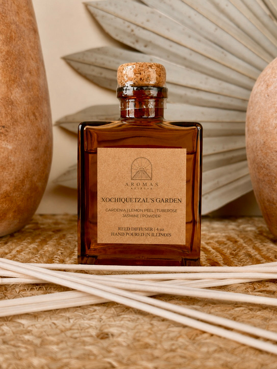 Aromas Aztecas Xochiquetzal's Garden reed diffuser in an amber glass bottle with natural reeds on a woven textured surface with terracotta pottery and dried palm leaf décor.