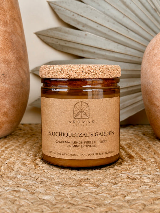 Aromas Aztecas Xochiquetzal's Garden soy candle in an amber glass vessel with a cork lid on a woven textured surface with terracotta pottery and dried palm leaf décor.