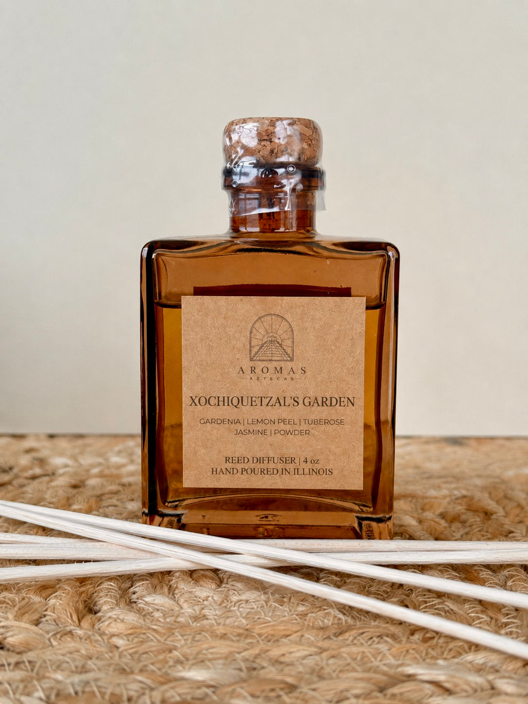 Aromas Aztecas Xochiquetzal's Garden reed diffuser in an amber glass bottle with natural reeds on a woven textured surface.
