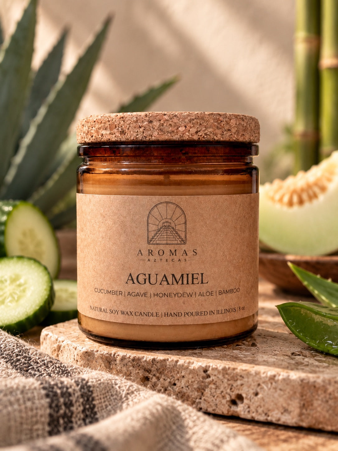 Aromas Aztecas Aguamiel soy candle with a cork lid on a rustic stone surface, surrounded by subtle cucumber, honeydew, aloe, agave, and bamboo accents in a warm, natural setting.