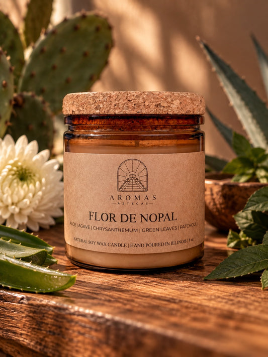 Aromas Aztecas Flor de Nopal candle in an amber glass jar with a cork lid, displayed on a warm wooden table with subtle aloe, agave, chrysanthemum, and green foliage surrounding it in soft golden light.