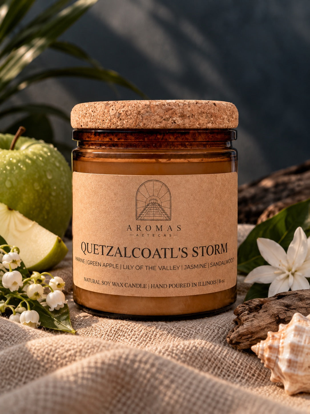Aromas Aztecas Quetzalcoatl’s Storm candle in an amber glass jar with a cork lid, styled on textured beige fabric against a dark, moody backdrop. Green apple, white flowers, leafy greenery, driftwood, and a seashell subtly reflect the candle’s marine, green apple, lily of the valley, jasmine, and sandalwood fragrance notes.
