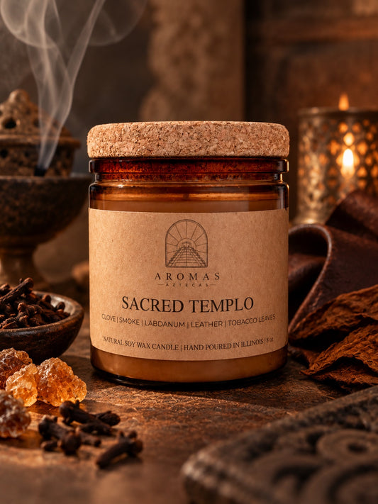 Aromas Aztecas Sacred Templo candle in an amber glass jar with a cork lid, styled in a warm, moody setting with cloves, labdanum resin, leather, tobacco leaves, wood, and subtle incense smoke. 