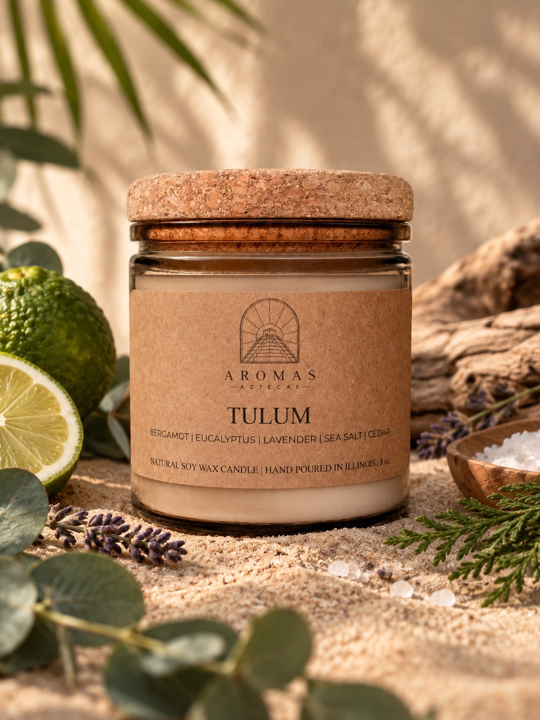 Aromas Aztecas Tulum candle in an amber glass jar with a cork lid, displayed on a sandy surface with eucalyptus leaves, lavender, lime, sea salt, and cedar accents. Warm natural lighting and a soft tropical backdrop create a subtle coastal setting.