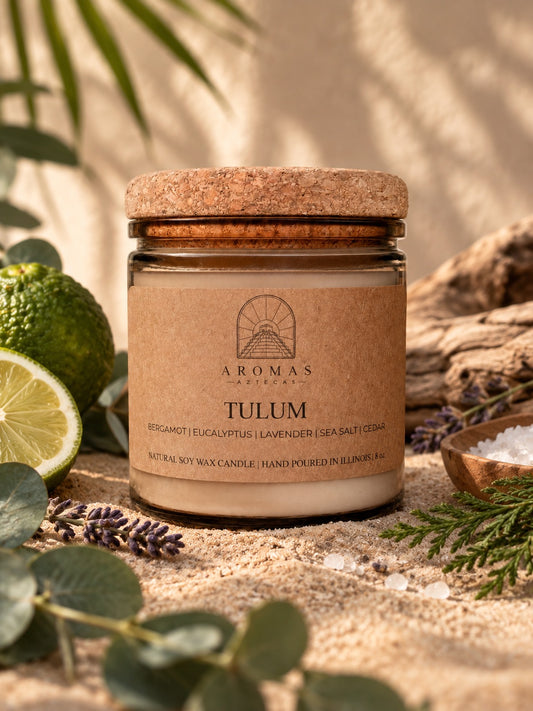 Aromas Aztecas Tulum candle in an amber glass jar with a cork lid, displayed on a sandy surface with eucalyptus leaves, lavender, lime, sea salt, and cedar accents. Warm natural lighting and a soft tropical backdrop create a subtle coastal setting.