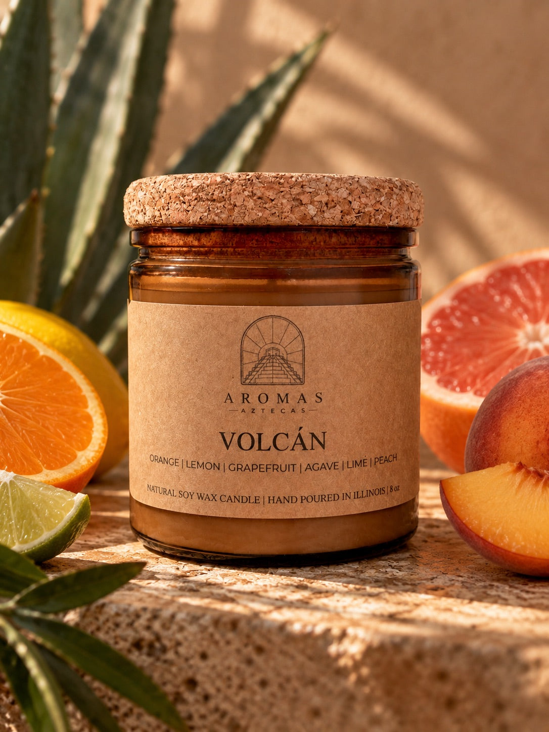 Aromas Aztecas Volcán candle in an amber glass jar with a cork lid, displayed on a warm textured surface with orange, grapefruit, lime, and peach slices and subtle agave leaves in the background.