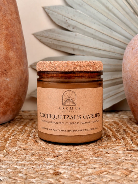 Aromas Aztecas Xochiquetzal's Garden soy candle in an amber glass vessel with a cork lid on a woven textured surface with terracotta pottery and dried palm leaf décor.
