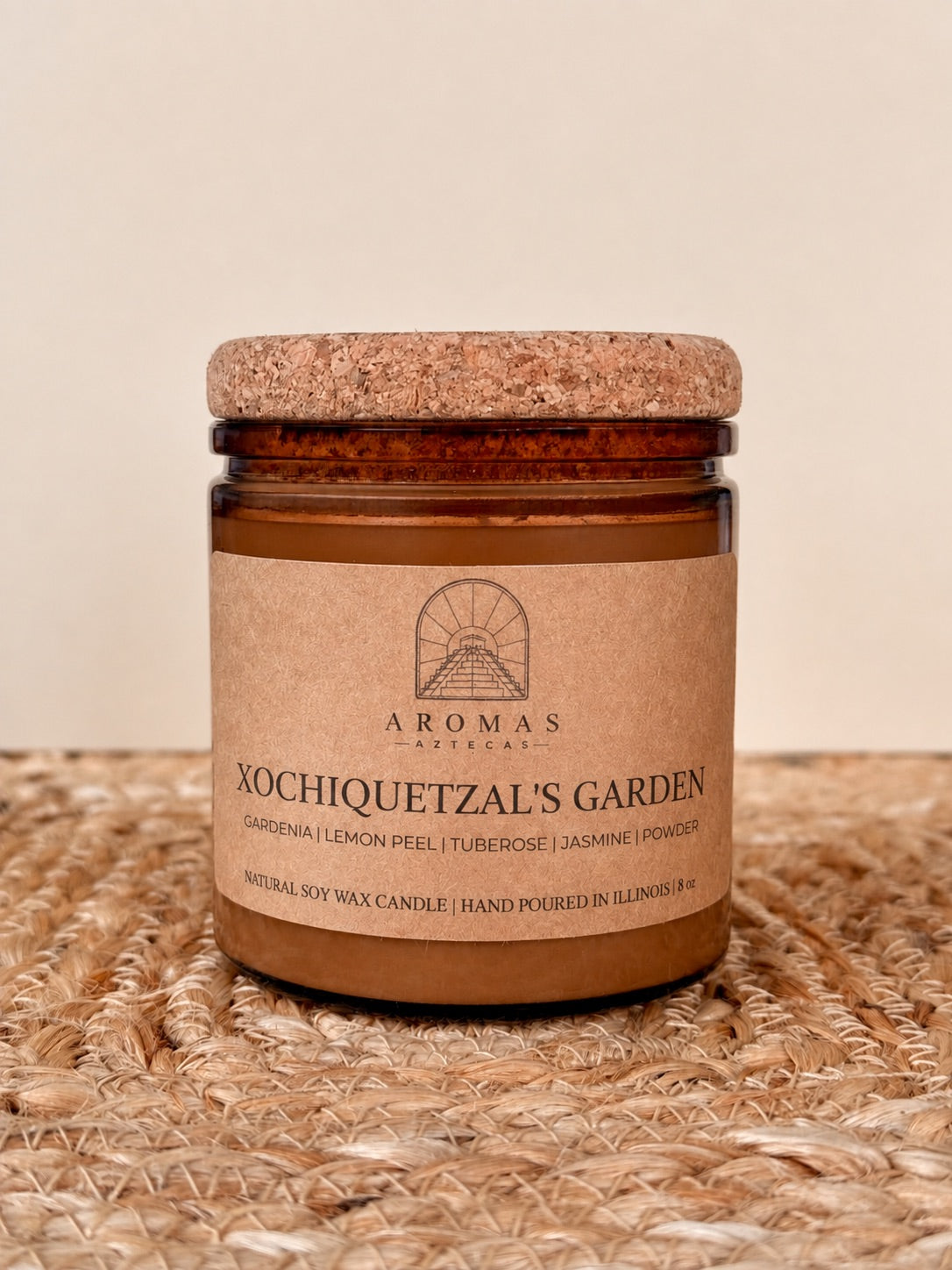 Aromas Aztecas Xochiquetzal's Garden soy candle in an amber glass vessel with a cork lid on a woven textured surface.