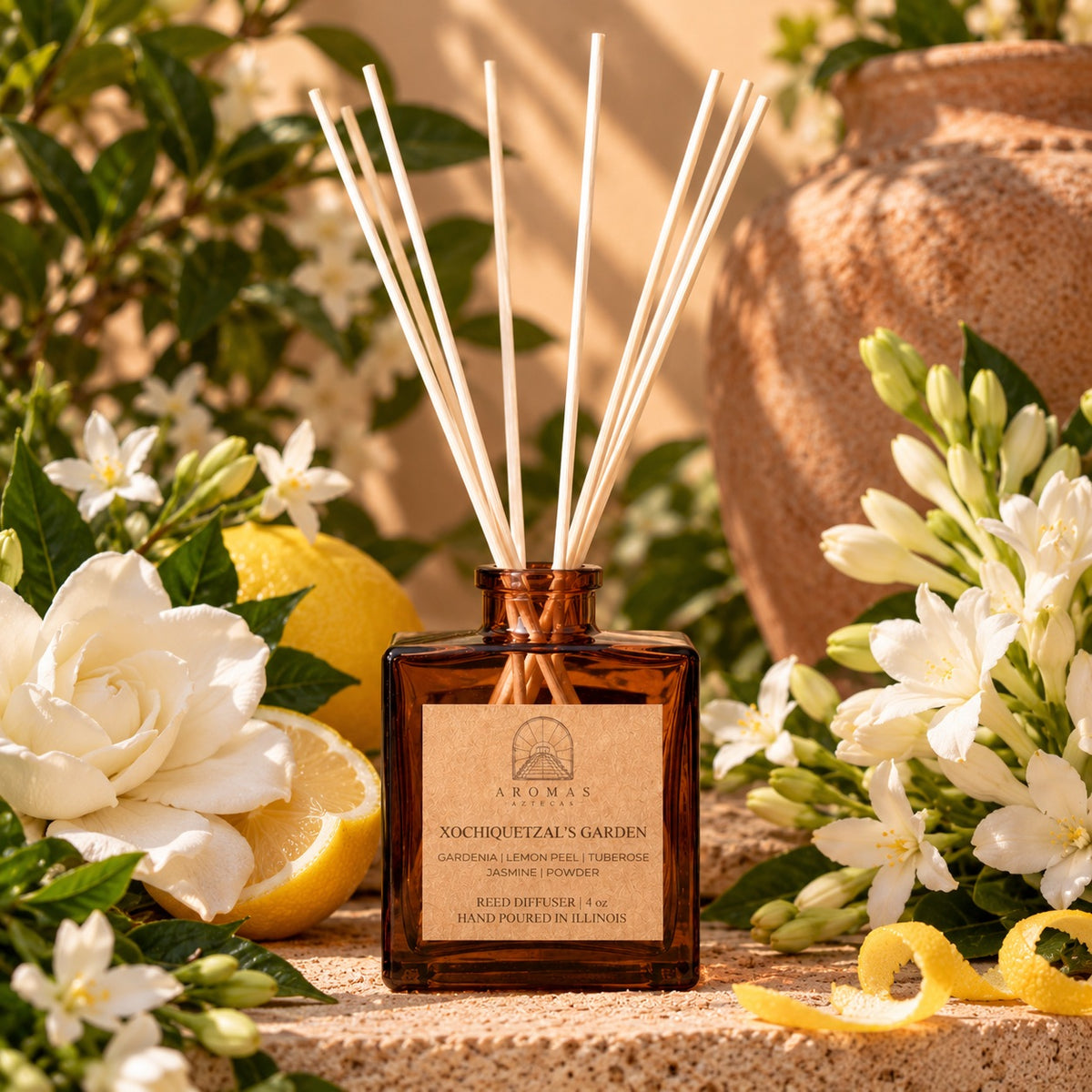 Aromas Aztecas Xochiquetzal’s Garden reed diffuser in an amber glass bottle with natural reeds, surrounded by gardenia, lemon, tuberose, jasmine, greenery, and warm terracotta accents against a sunlit background.