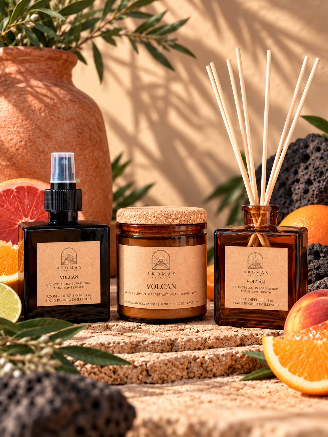 Aromas Aztecas Volcán fragrance collection featuring an amber glass room + linen spray, amber glass soy candle, and amber glass reed diffuser with natural reeds inserted, styled with oranges, lemons, grapefruit, agave, lime, and peach accents on a natural stone surface against a sunlit background.