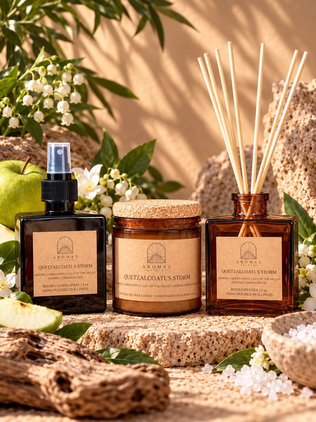 Aromas Aztecas Quetzalcoatl’s Storm fragrance collection featuring an amber glass room + linen spray, amber glass soy candle, and amber glass reed diffuser with natural reeds inserted, styled with green apple, marine-inspired elements, white florals, and natural stone accents against a sunlit background.
