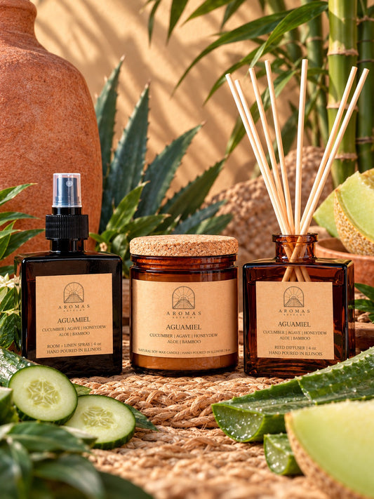 Aromas Aztecas Aguamiel fragrance collection featuring an amber glass room + linen spray, amber glass soy candle, and amber glass reed diffuser with natural reeds inserted, styled with cucumber, aloe, honeydew, agave, and bamboo accents on a woven natural-fiber surface against a sunlit background.