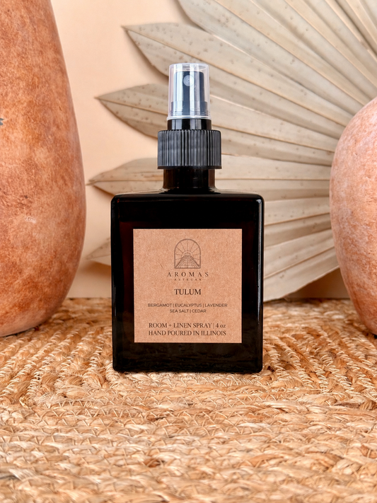 Aromas Aztecas Tulum room and linen spray in an amber glass bottle on a woven textured surface with terracotta pottery and dried palm leaf décor.