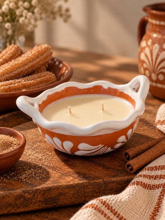 Churros handcrafted terracotta talavera cazuelita candle with two wicks on a rustic wooden table, surrounded by churros, cinnamon sticks, terracotta pottery, and a woven textile.