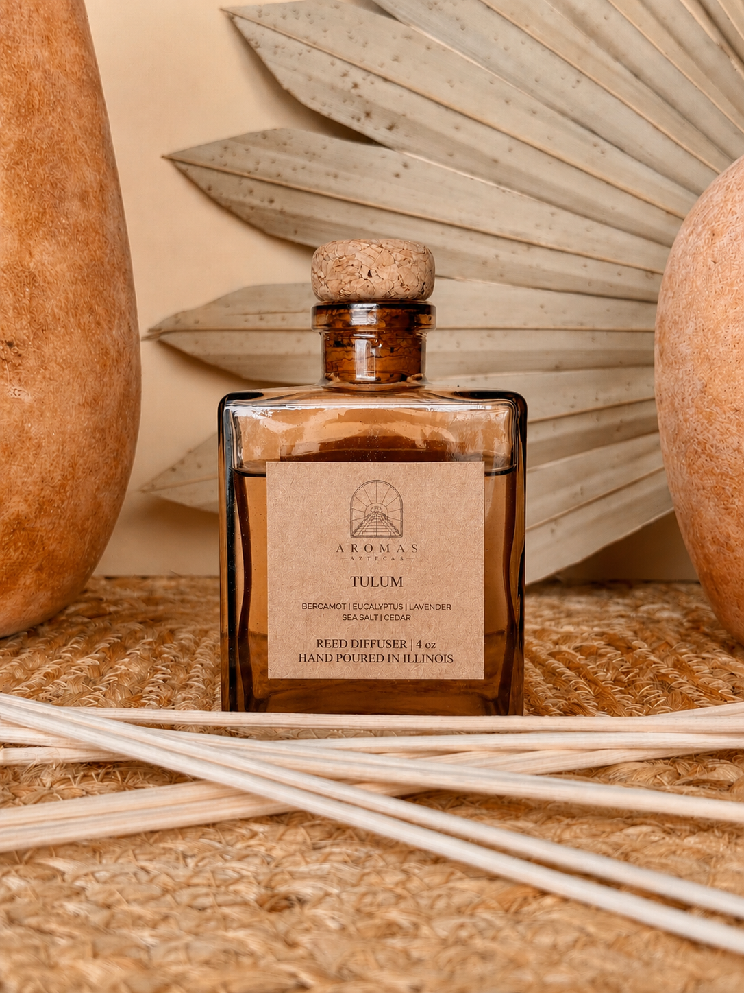 Aromas Aztecas Tulum reed diffuser in an amber glass bottle with natural reeds on a woven textured surface with terracotta pottery and dried palm leaf décor.