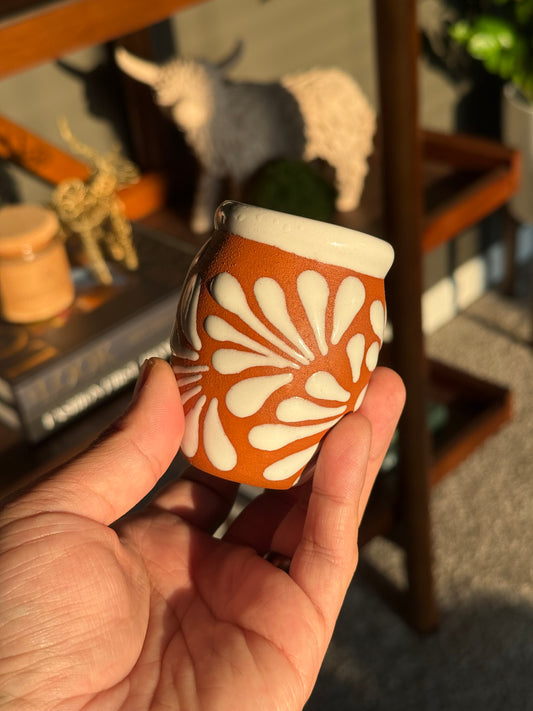 Hand holding a small terracotta colored ceramic Cempasúchil velita with a white floral pattern and white rim, photographed in warm natural light with a blurred bookshelf in the background.
