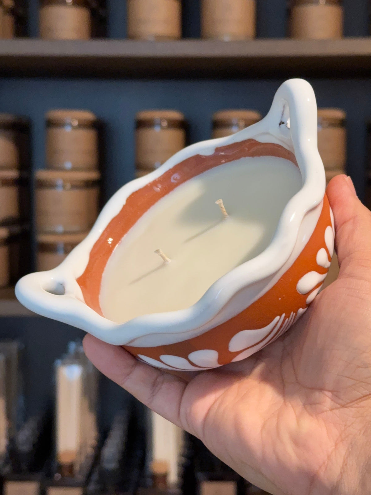 Handcrafted terracotta talavera cazuelita candle with two wicks, held in a hand against a softly blurred shelf of candle vessels.