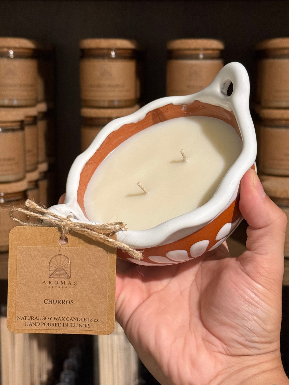 Churros cazuelita candle with two wicks in a terracotta artisan talavera vessel, held in a hand with a branded product tag, photographed against shelves of candle jars.