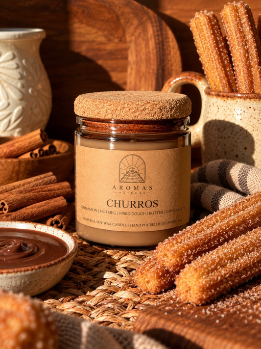 Aromas Aztecas Churros soy candle in an amber glass vessel with a cork lid on a woven textured surface next to cinnamon sticks, a bowl of chocolate and surrounded by churros.