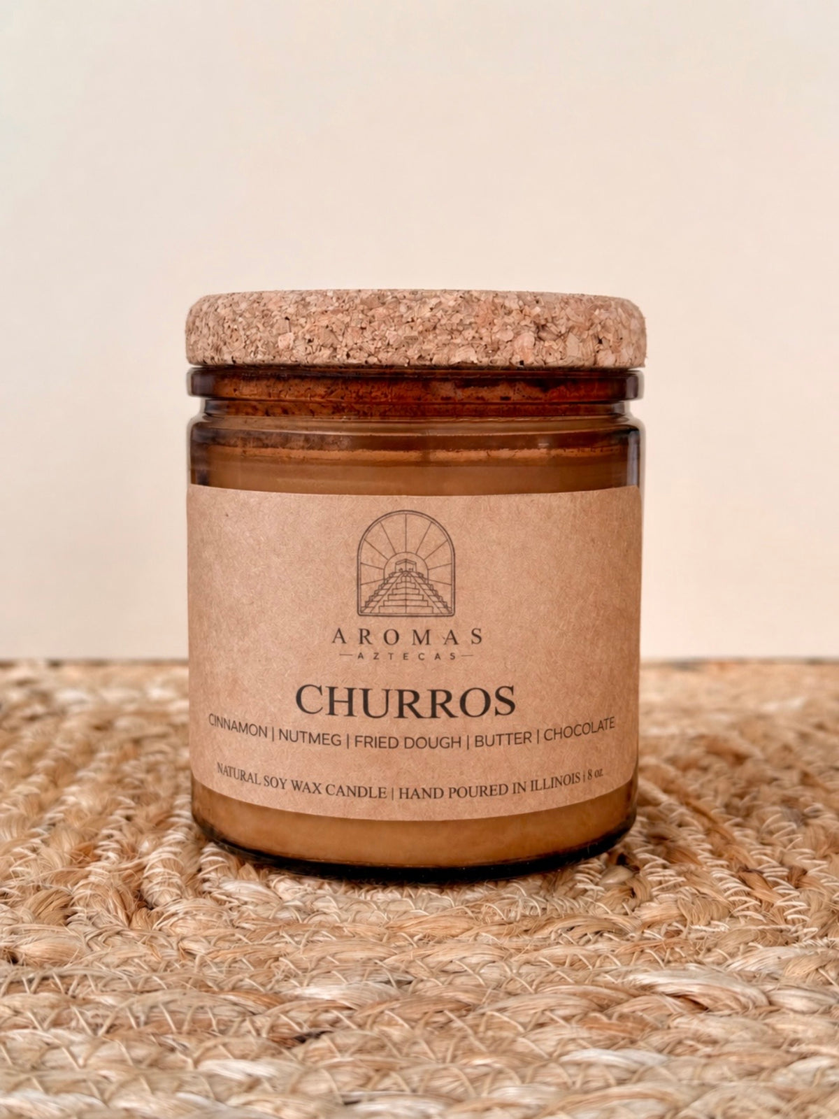 Aromas Aztecas Churros soy candle in an amber glass vessel with a cork lid on a woven textured surface.