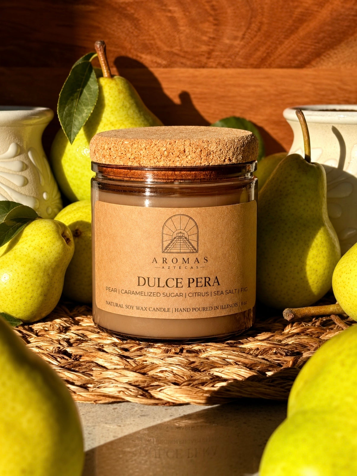 Aromas Aztecas Dulce Pera soy candle in an amber glass vessel with a cork lid on a woven textured surface surrounded by green pears against a wooden grain background.