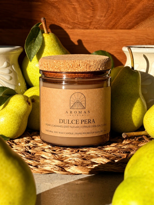 Aromas Aztecas Dulce Pera soy candle in an amber glass vessel with a cork lid on a woven textured surface surrounded by green pears against a wooden grain background.