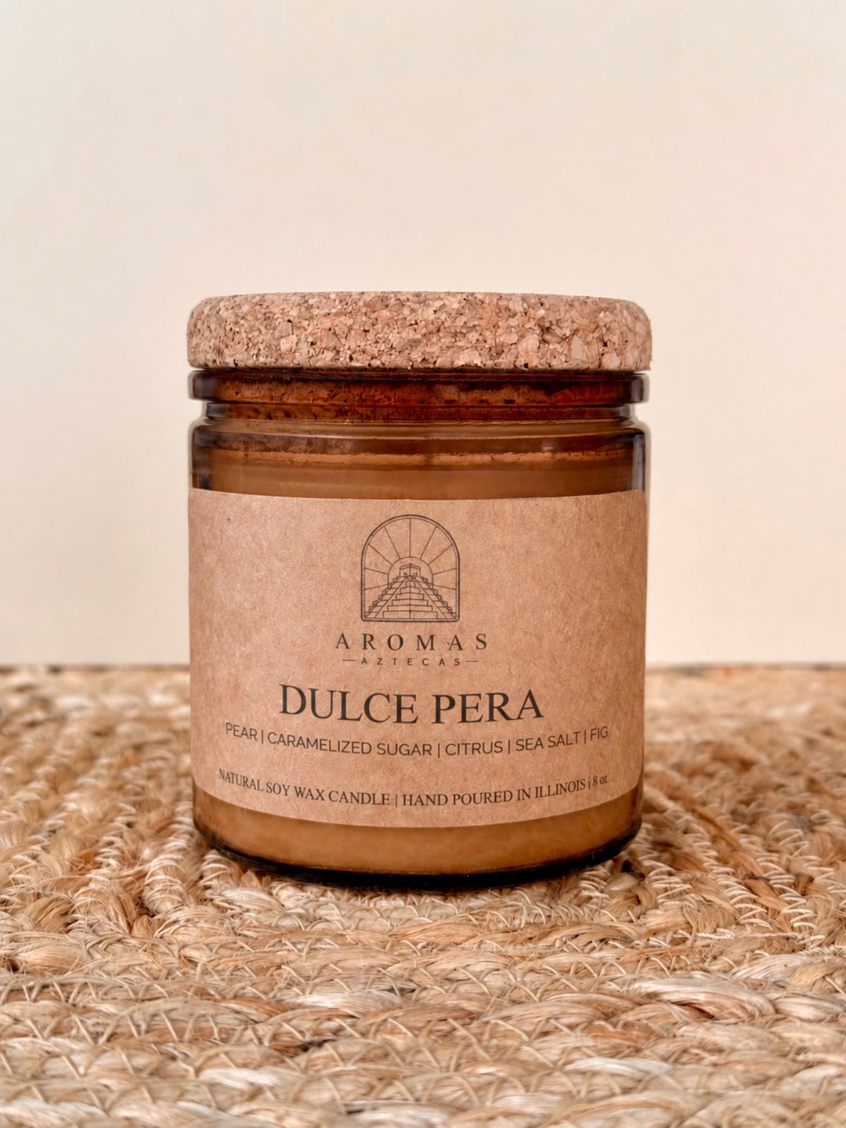 Aromas Aztecas Dulce Pera soy candle in an amber glass vessel with a cork lid on a woven textured surface.