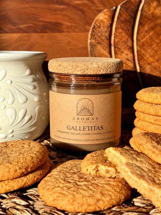 Aromas Aztecas Galletitas soy candle in an amber glass vessel with a cork lid on a woven textured surface next to cinnamon sugar cookies and a white talavera mug against a wooden grain background.