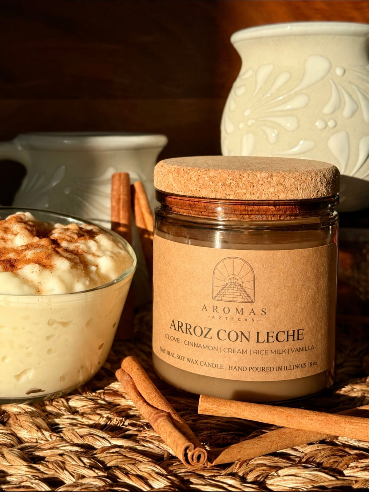 Aromas Aztecas Arroz con Leche soy candle in an amber glass vessel with a cork lid on a woven textured surface behind cinnamon sticks and next to a cup of rice pudding.
