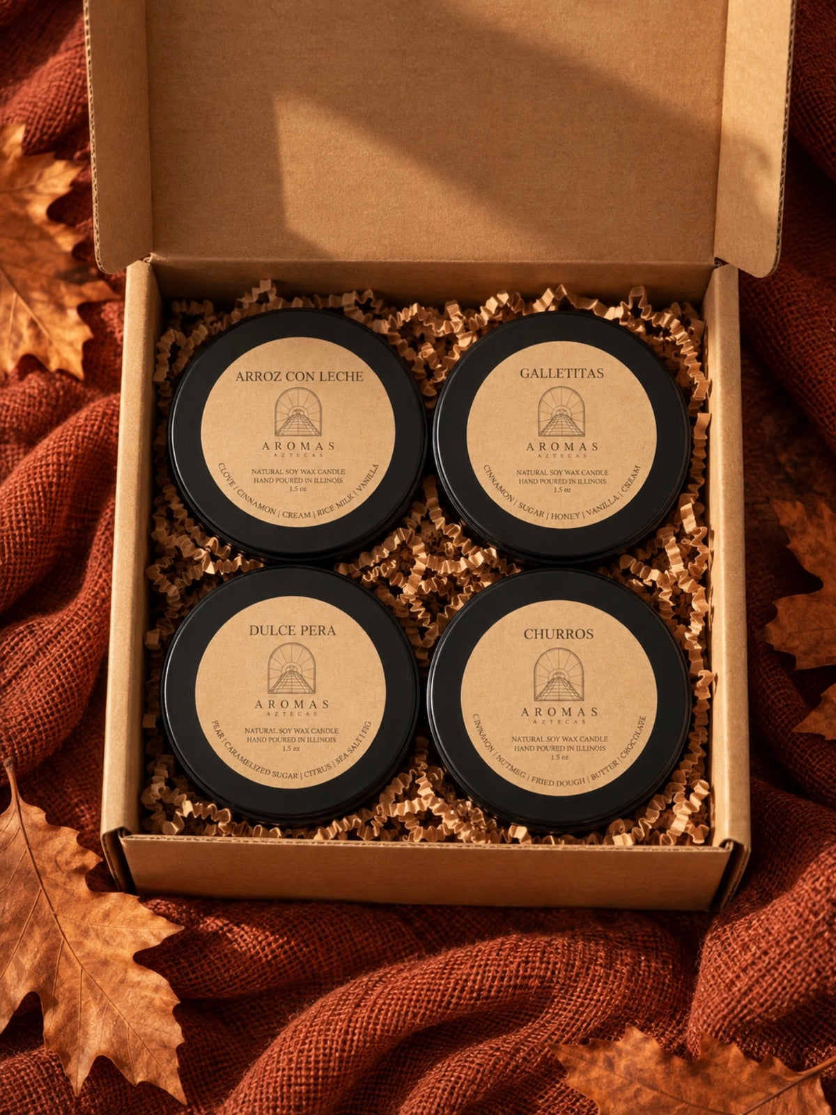 Open kraft gift box containing four Aromas Aztecas 1.5 oz fall discovery candles: Arroz con Leche, Galletitas, Dulce Pera, and Churros, nestled in kraft crinkle paper. The box is surrounded by terracotta fabric and autumn leaves.